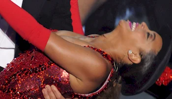 A woman in a sparkling red outfit and long red gloves leans back, smiling widely, while being held up by another person. She wears a black hat with a red flower.