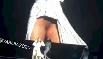 A performer in a white outfit sings on stage with a large screen showing a close-up behind her. There are backup dancers and the audience is visible in the dark foreground.