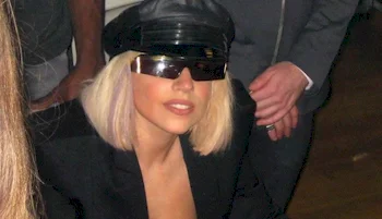 Lady Gaga with blonde hair wearing a black hat, sunglasses, and a black outfit sits indoors. Other people are standing nearby, partially visible.