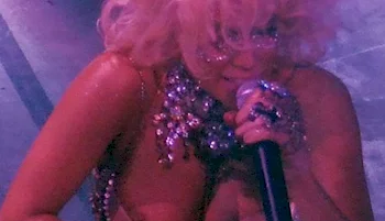 A woman with blonde, curly hair and glittery makeup sings into a microphone on stage, wearing a sparkling outfit with elaborate jewelry, under purple lighting.