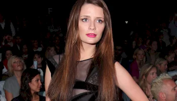 A woman with long brown hair and a sheer black dress stands confidently in front of a seated crowd at what appears to be a fashion event.