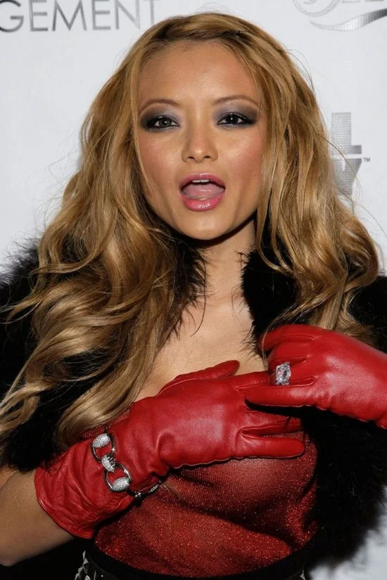 A woman with long wavy blonde hair wears red gloves, a black fur stole, and a red outfit. She is showing off a ring on her gloved hand and has her mouth slightly open.