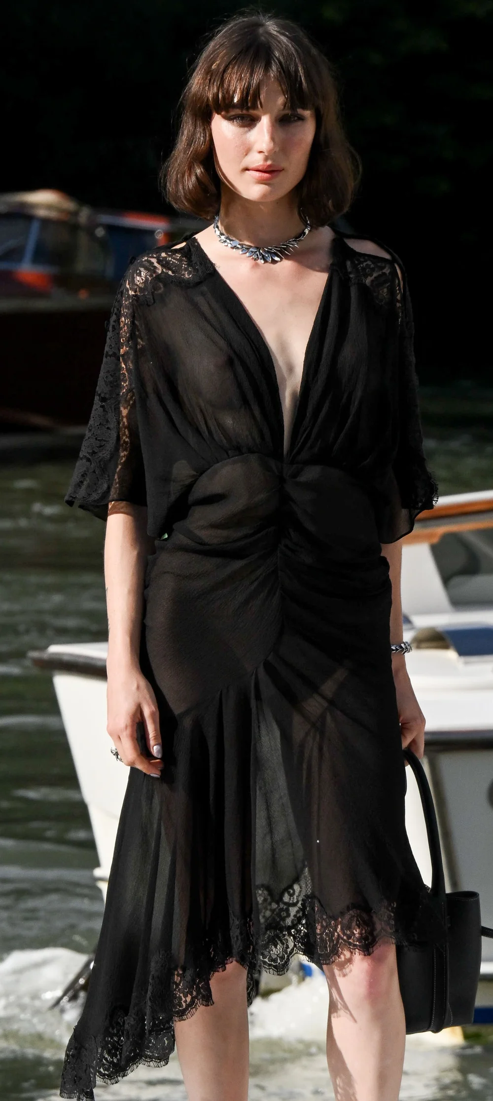 Alice-Pagani-See-Through-83rdVFF1 A woman in a sheer black dress with lace details stands outdoors near water and boats. She has short brown hair, wears a silver necklace, and holds a black handbag.