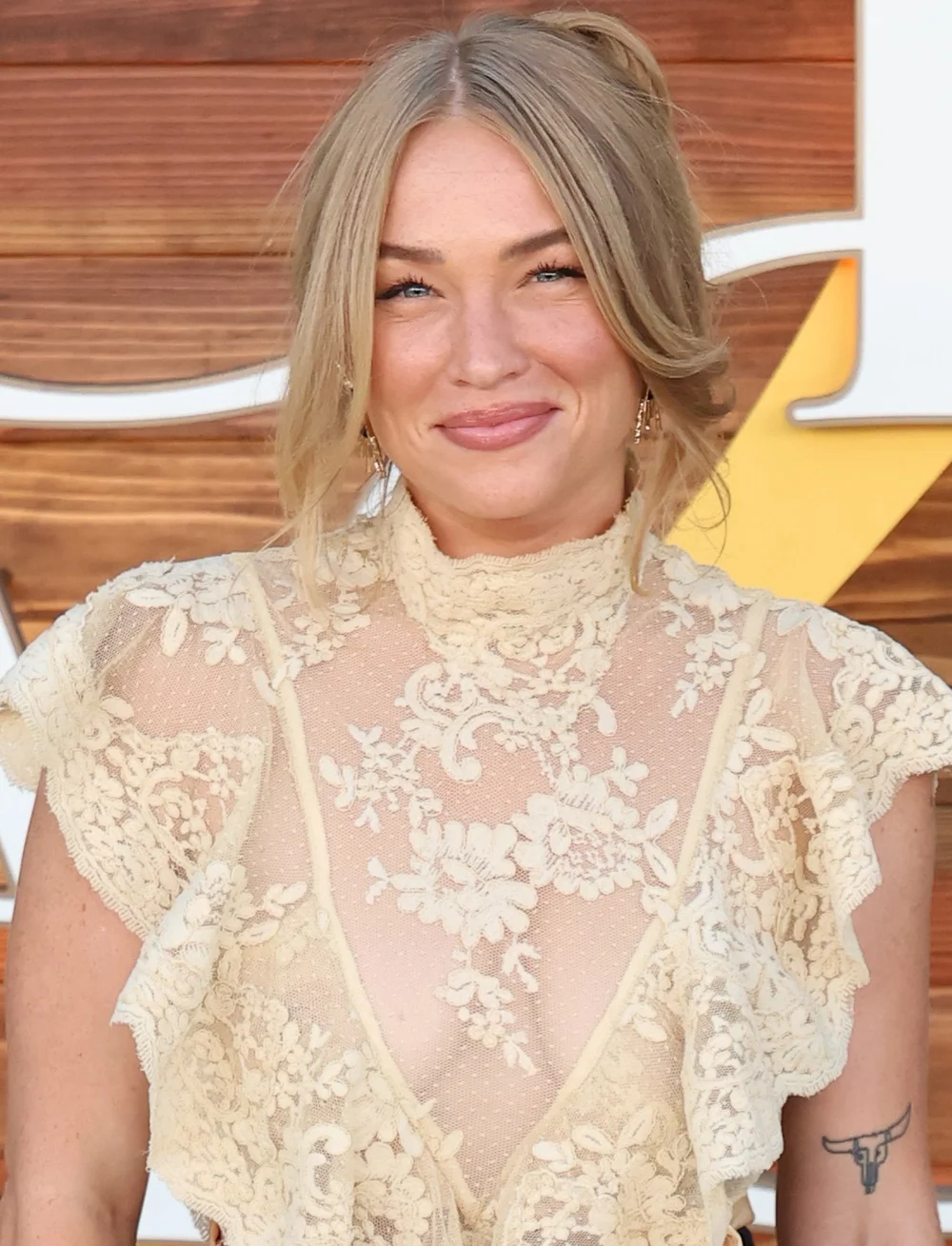 A woman with blonde hair in a loose updo wears a sheer, lacy cream blouse and smiles at the camera. The background features a wooden wall with a partial yellow and white sign.