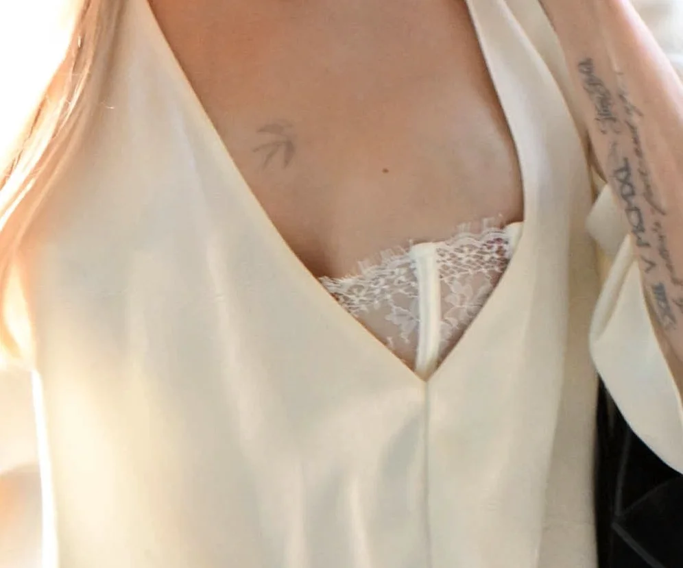 AngelinaJolieNipJFKAirportPic2 A close-up of a person wearing a cream-colored top with a deep V-neck, revealing a white lace undergarment. A faint tattoo is visible on their chest and script tattoos on their arm.