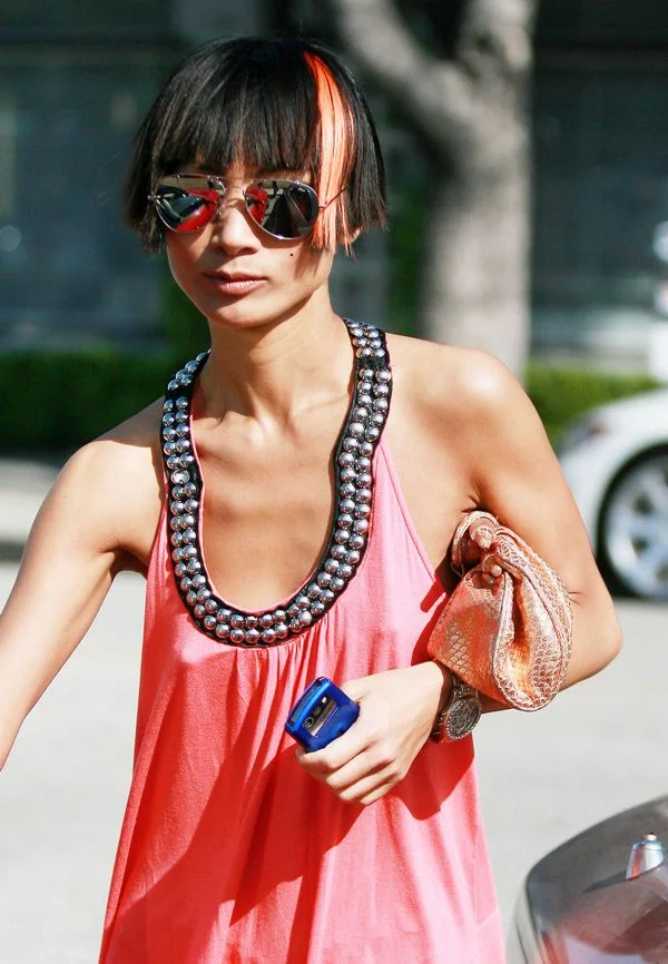 A person wearing sunglasses and a pink dress with metallic trim holds a shiny clutch and a blue phone, with short black hair featuring an orange streak.