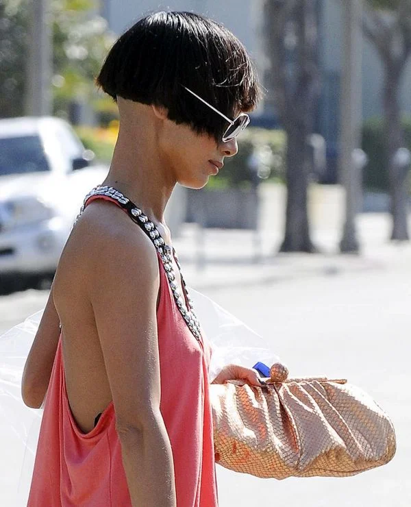 Person wearing sunglasses and a sleeveless pink top, holding a textured peach-colored bag. Their hair is cut in a dramatic bowl style with a sharp line above the ears and nape, exposing the neck.