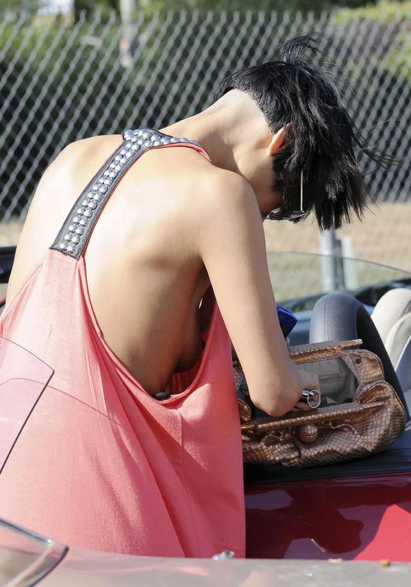 A person in a loose pink top leans into a car, facing away, with short black hair and sunglasses. A tan handbag rests on the car seat beside them. A chain-link fence and greenery are in the background.