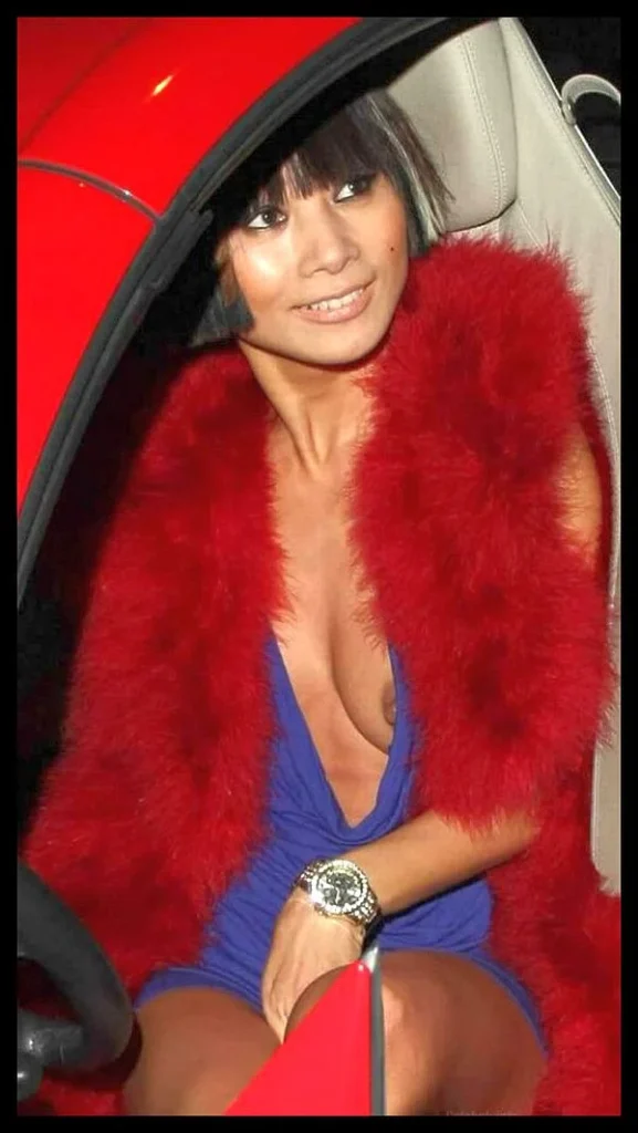 A woman wearing a red fur stole and blue dress sits in a car, smiling.