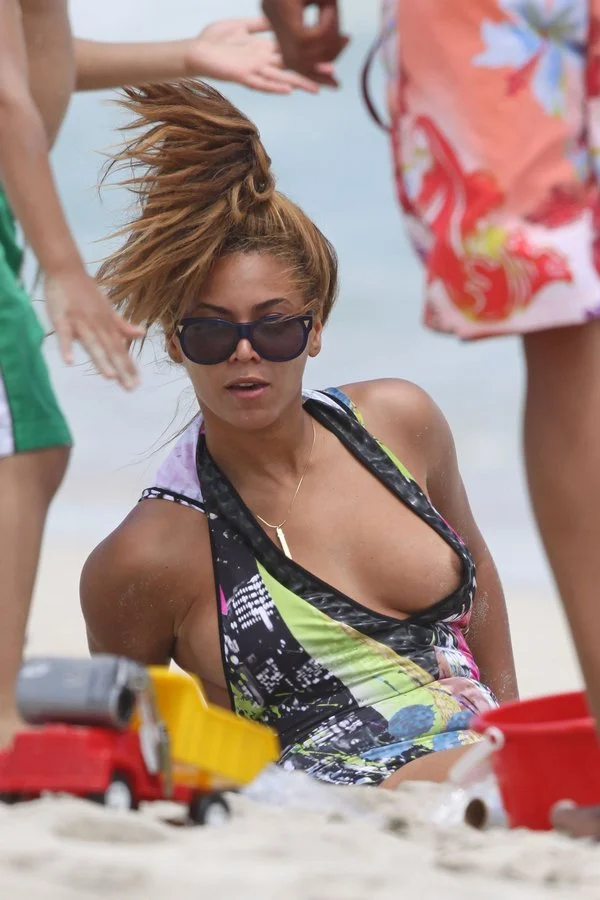Beyonce wearing sunglasses and a swimsuit sits on a sandy beach near toy trucks and buckets, with children playing nearby. Her hair is partially up and she appears surprised or reacting to something.