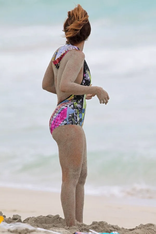 A woman with sandy skin stands on a beach wearing a colorful one-piece swimsuit, looking toward the ocean with her hair up. The background shows waves and a cloudy sky.