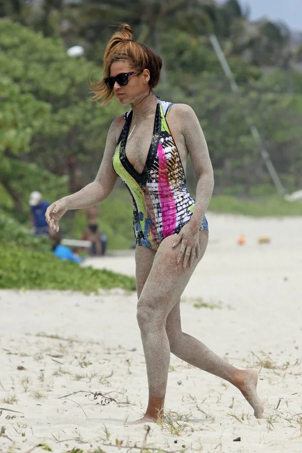 Beyonce wearing sunglasses and a colorful swimsuit walks on a sandy beach, with sand covering her arms and legs. Green foliage and blurred people are visible in the background.
