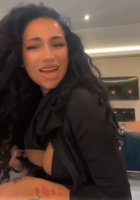 A woman with long curly hair, wearing a black top, smiles while sitting indoors. The background shows a modern room with a lighted ceiling and wall-mounted screens.