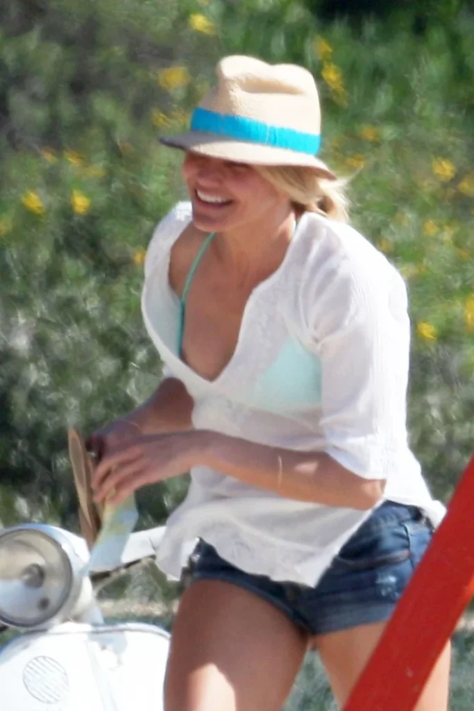 A woman wearing a sunhat, white blouse, and denim shorts smiles outdoors near a white scooter, with greenery and yellow flowers in the background 1/3.
