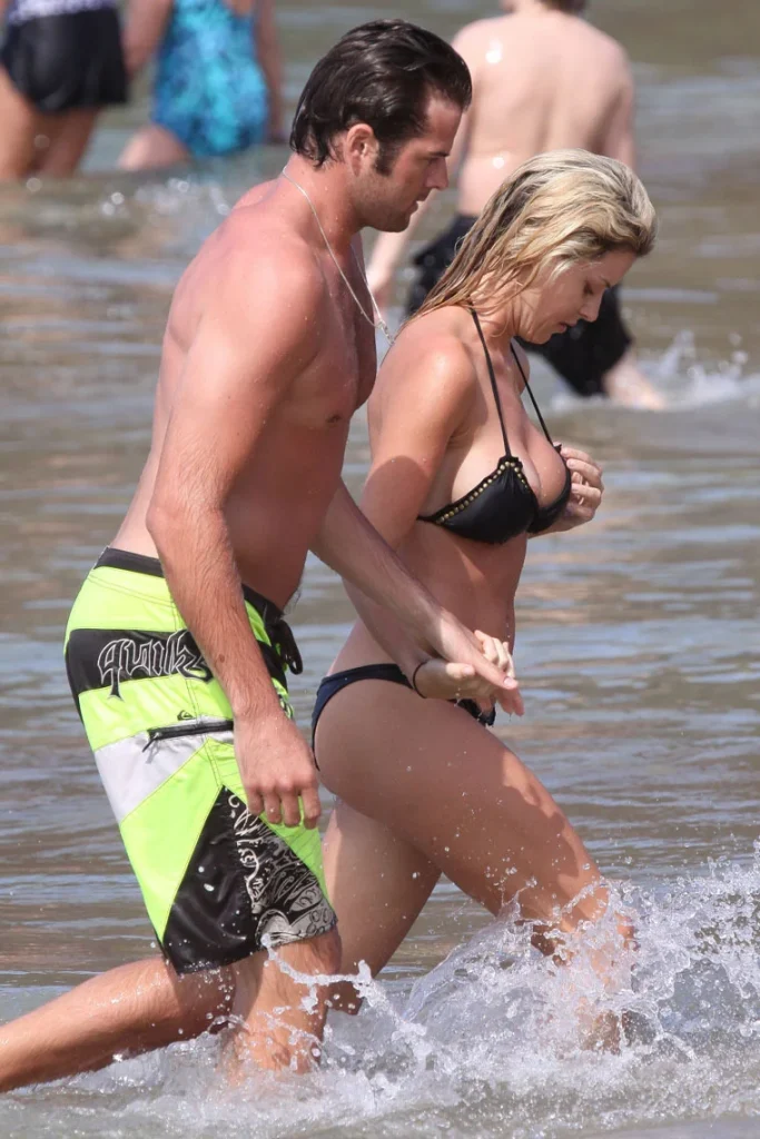 A man in green swim trunks and a woman in a black bikini walk hand in hand through shallow ocean water at a crowded beach.
