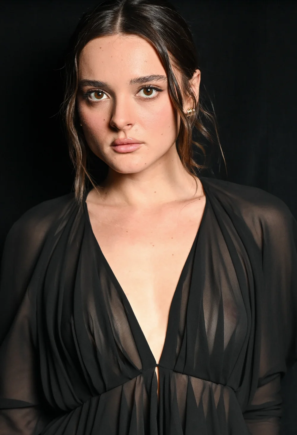 CharlotteLawrenceSeeThroughKhaite26NYFWPic A woman with long brown hair in loose waves wears a sheer black dress with a deep V-neckline, standing against a black background and looking directly at the camera with a neutral expression.