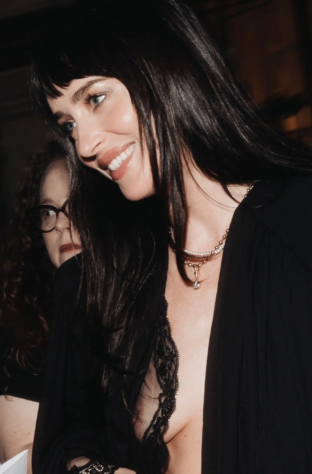 DakotaJohnsonNipSlipVOGUEWorld2026MilanPic1 Woman with long dark hair, wearing a black lace-trimmed outfit and layered necklaces, smiling. Another person with glasses is partially visible in the background.
