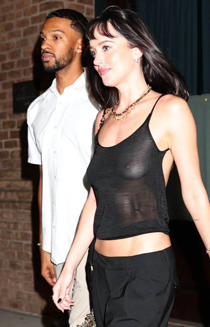 DakotaJohnsonSheerTopTWAptPic1 A woman in a sheer black tank top and black pants walks beside a man in a short-sleeve white shirt outdoors at night, with a brick wall in the background.