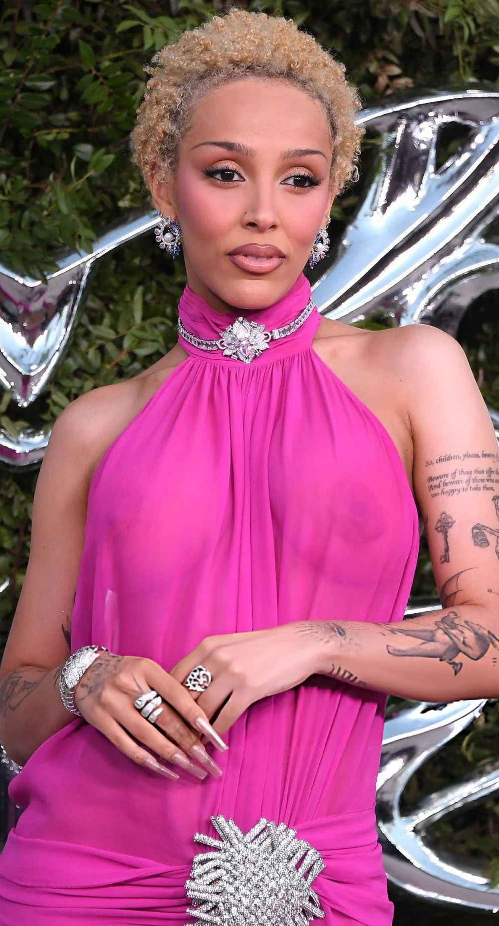 DojaCatSeeThroughLucasMuseumOpeningPic1 A woman with short curly blond hair wears a bright pink halter dress, silver jewelry, and rings, posing in front of a silver abstract background with tattoos visible on their arms.