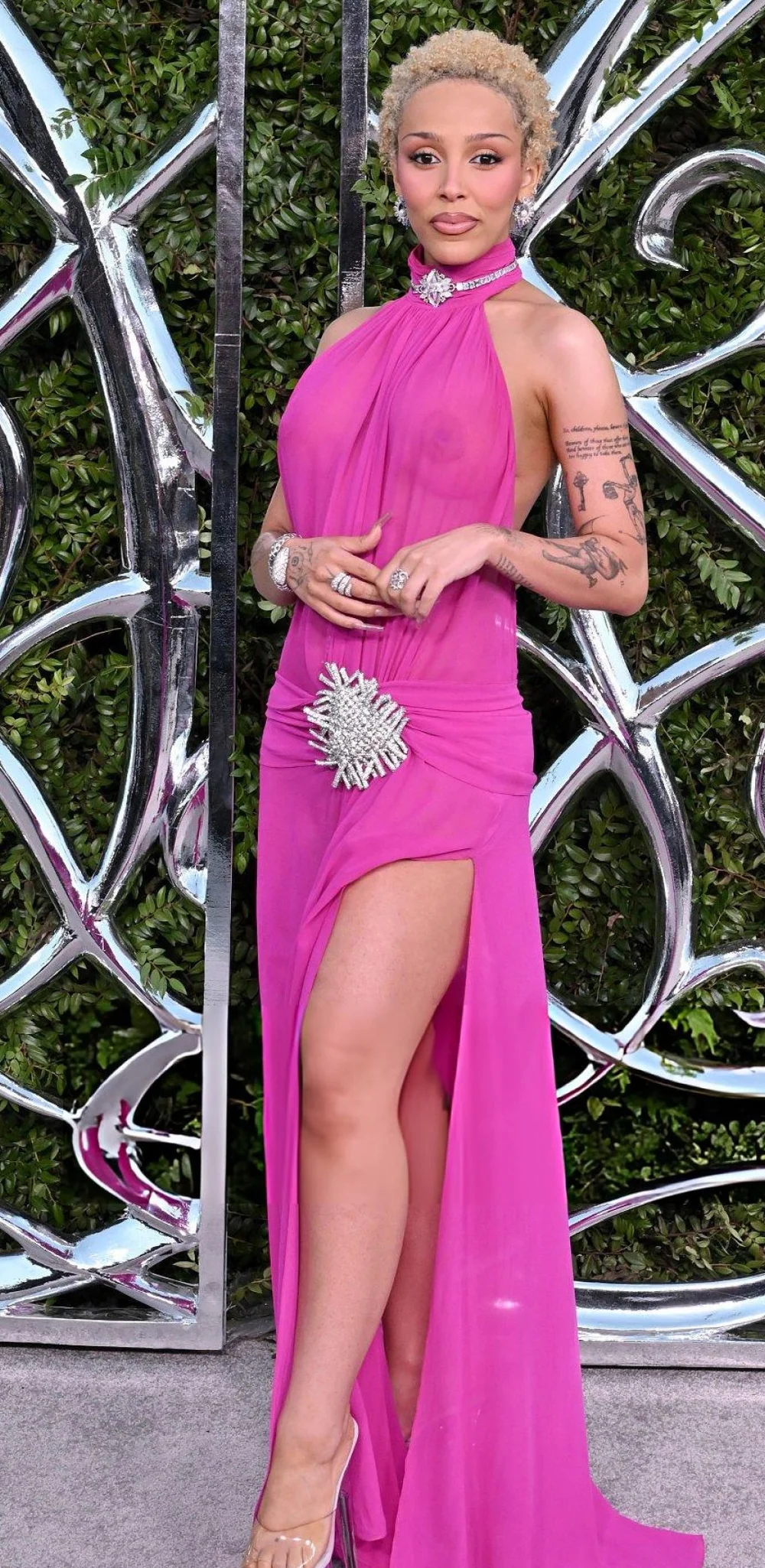 DojaCatSeeThroughLucasMuseumOpeningPic2 A woman wearing a sheer hot pink gown with a high slit, silver starburst belt, and jewelry stands in front of a metallic abstract background with greenery visible behind the structure.