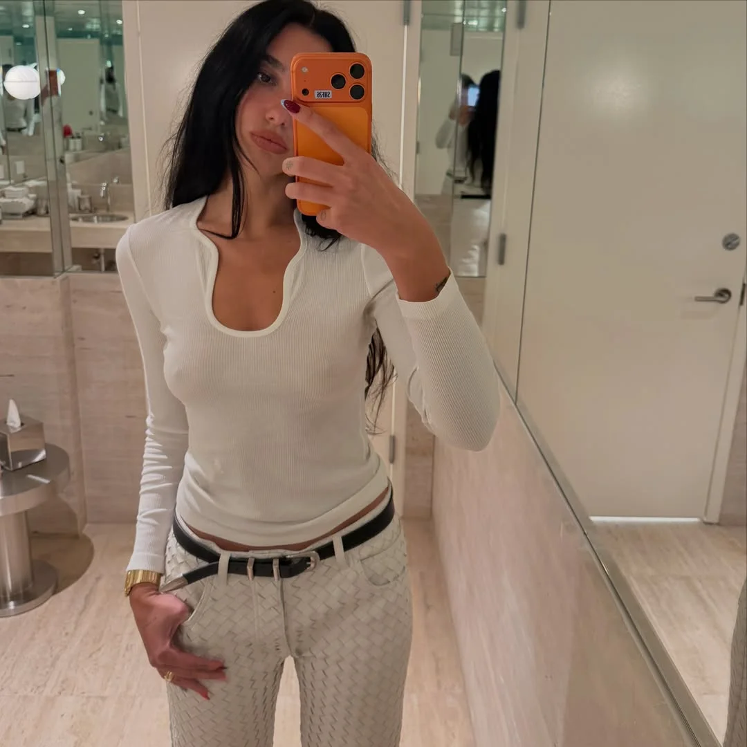 DuaLipaBralessSelfieAllWhite A woman with long dark hair takes a mirror selfie in a bathroom. They wear a white long-sleeve top and patterned white pants, holding an orange phone. The bathroom features beige walls and visible sinks.
