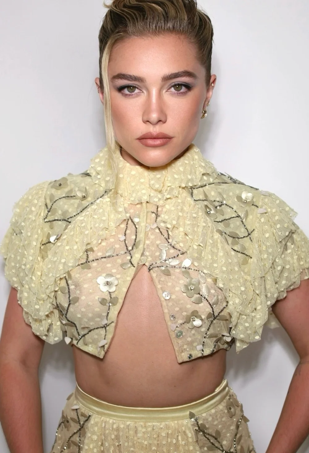 FlorencePughSheerEastOfEdenScreeningPic A woman with blonde hair styled up wears a sheer, pale yellow ruffled top with sequin details and a keyhole cutout, standing against a plain white background.