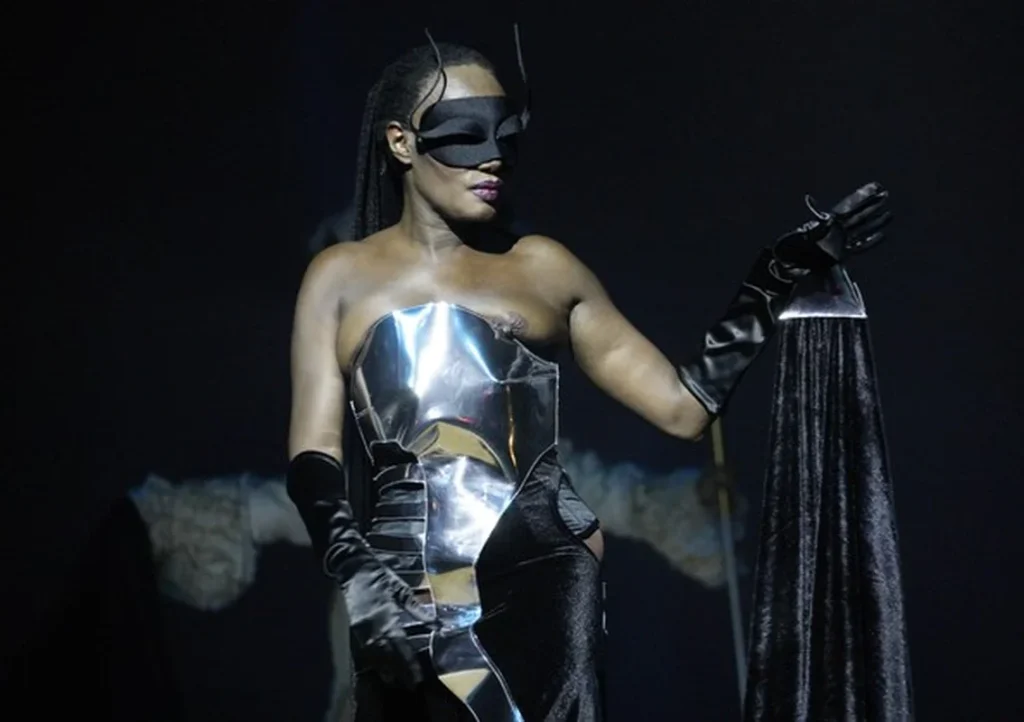 Grace Jones wearing a shiny, sculpted metallic corset, long black gloves, and a black mask stands on stage, holding a black velvet-like fabric. The background is dark and dramatic.