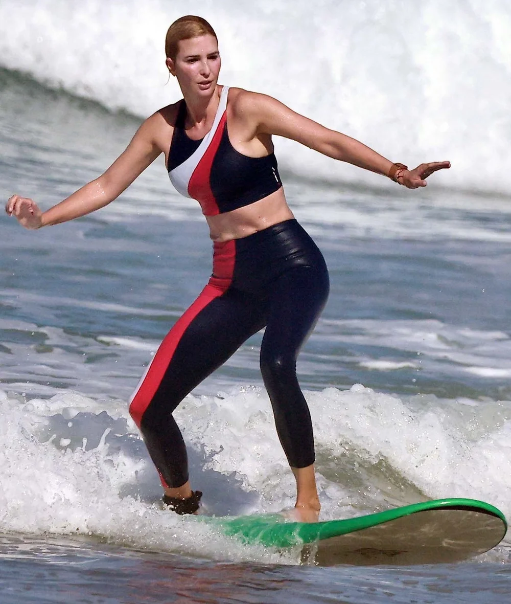 A woman in a black, red, and white athletic outfit surfs on a green surfboard, balancing on a small wave with the ocean and white surf behind her.