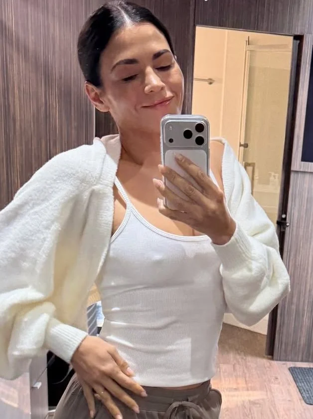 JennaDewanWhiteTankSweaterPokiesSelfiePic Woman taking a selfie in a bathroom mirror, wearing a white tank top and a white sweater draped over her shoulders, smiling slightly.