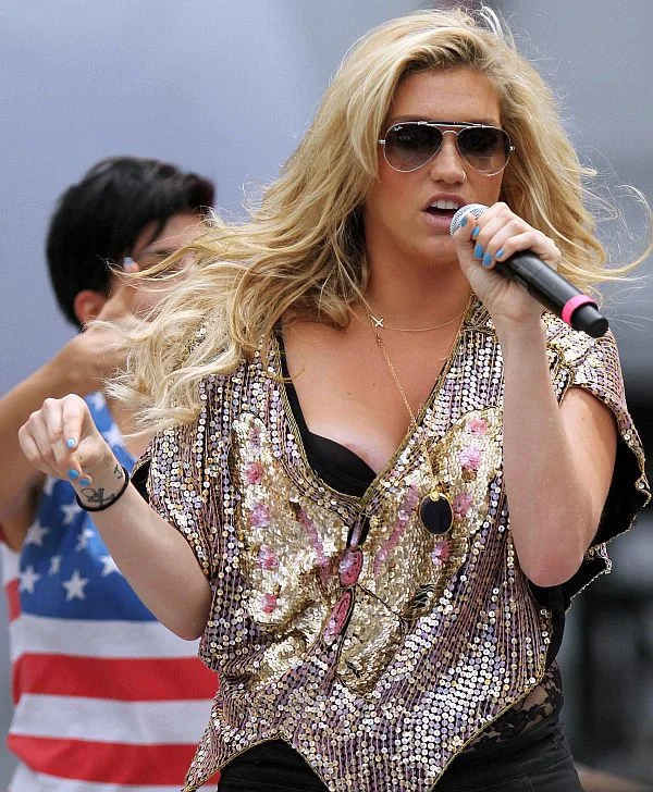 KeshaAreolaSlipUpskirtCanada1 A woman with long blonde hair and sunglasses sings into a microphone on stage, wearing a sequined top. Another person in the background wears a shirt with an American flag design.