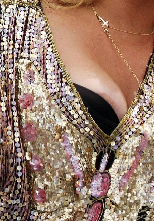 KeshaAreolaSlipUpskirtCanada2 A close-up of a woman's breast who is wearing a sequined, gold and pink outfit with a low neckline, a black inner garment, and a delicate gold necklace with a cross pendant.