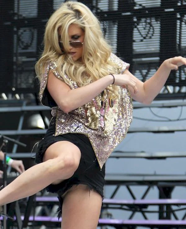 KeshaAreolaSlipUpskirtCanada3 A woman with long blonde hair, sunglasses, and a sparkly top dances energetically on stage, lifting one leg and raising her arms, with a blurred concert background behind her.