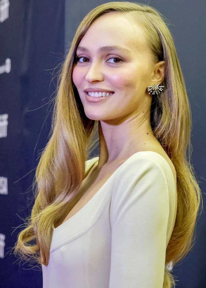 Lily-RoseDeppPokiesAlphaGang26TIFFPic1 A woman with long blonde hair and a white dress smiles at an event, wearing a large spiky earring. The background is dark and blurred, giving the photo a formal atmosphere.