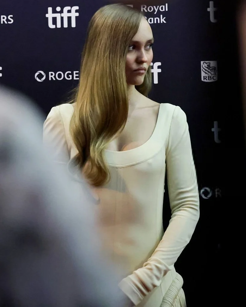 Lily-RoseDeppPokiesAlphaGang26TIFFPic2 A woman in a long-sleeved, cream-colored dress stands on a red carpet at TIFF, with her long, wavy hair draped over one shoulder, looking to the side.