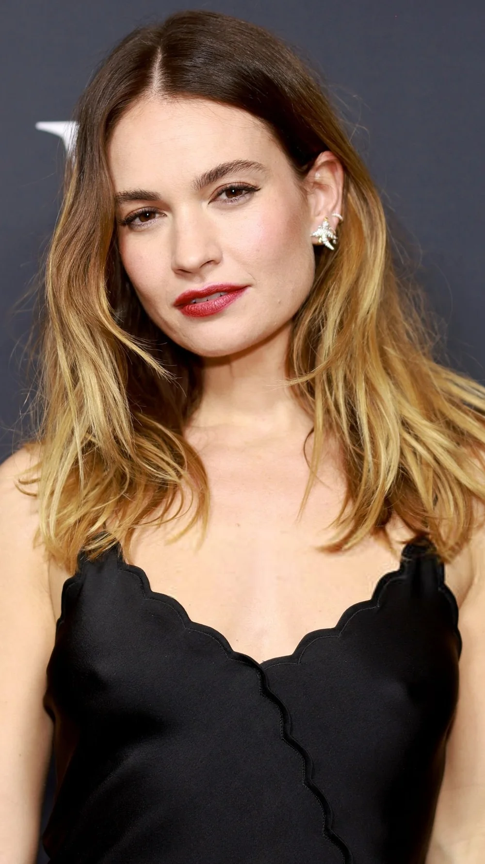 LilyJamesPokiesBadLieutenantTokyoTIFFPic A woman with wavy, light brown hair wearing a black scalloped dress and pearl earrings stands in front of a dark background, looking at the camera with a neutral expression.