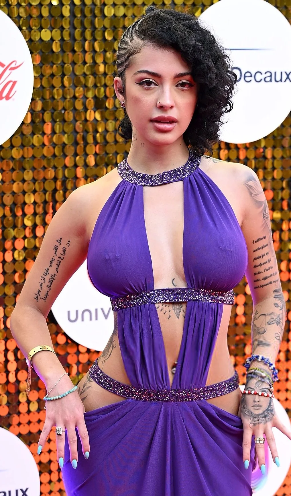 A woman poses on a red carpet wearing a purple cut-out dress, with tattoos on their arms and body, curly black hair styled to one side, and colorful painted nails. Gold sequin background and event logos are visible.