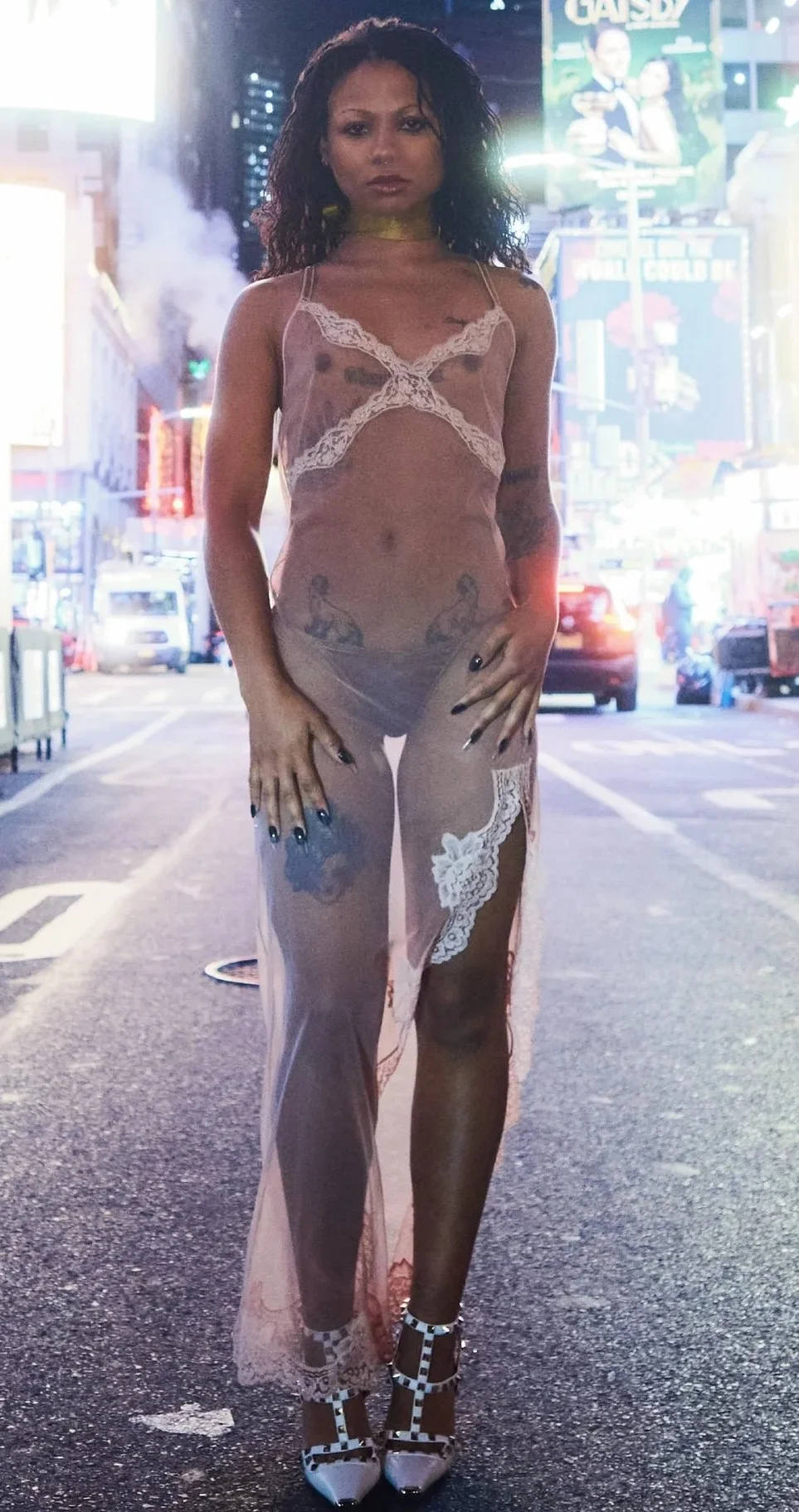 A woman stands confidently in the middle of a city street at night, wearing a sheer, lacy dress and heels. Bright city lights and cars are visible in the background.