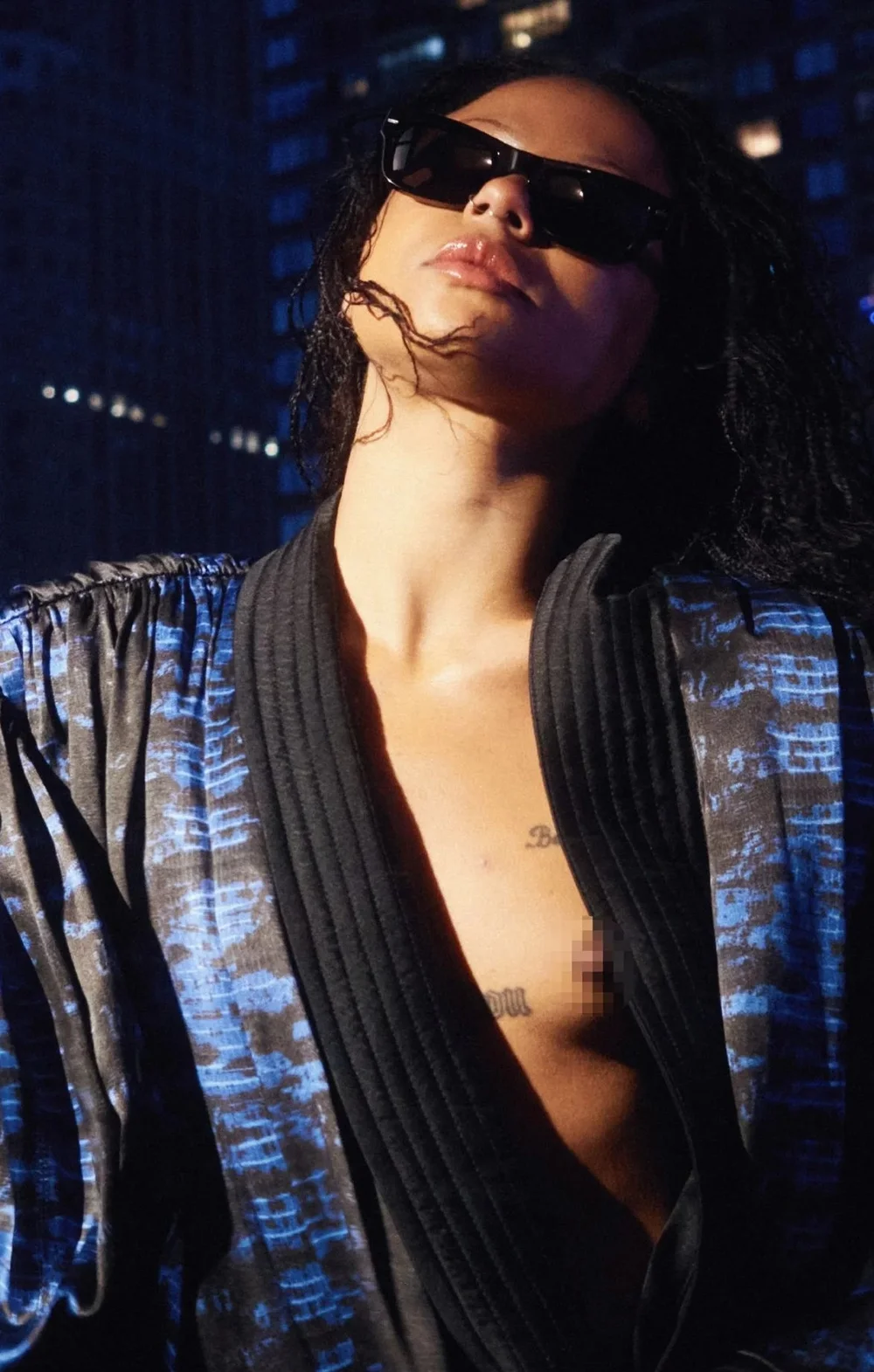 A woman wearing sunglasses and a blue patterned robe stands outdoors at night, with part of their chest exposed. City lights and tall buildings are visible in the dark background.