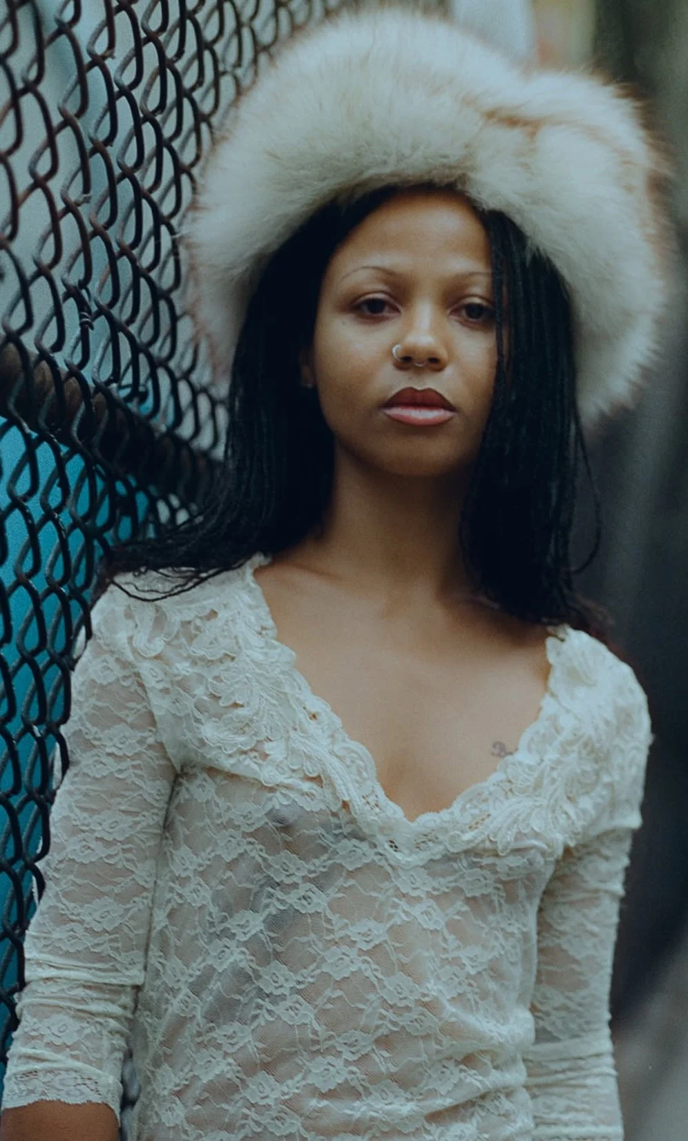 MyhalaSeeThroughElektraPic1 Woman with dark hair and a large white fur hat standing against a chain-link fence, wearing a sheer white lace long-sleeve top.