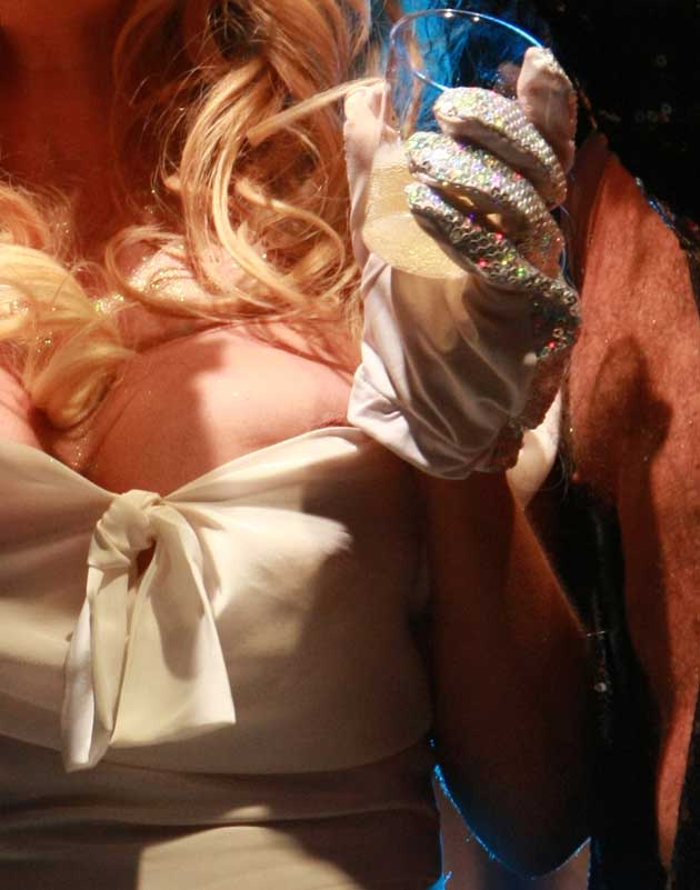A person in a white dress with long blonde hair holds a drink, wearing sparkly gloves and white satin gloves, with part of another persons arm visible beside them.