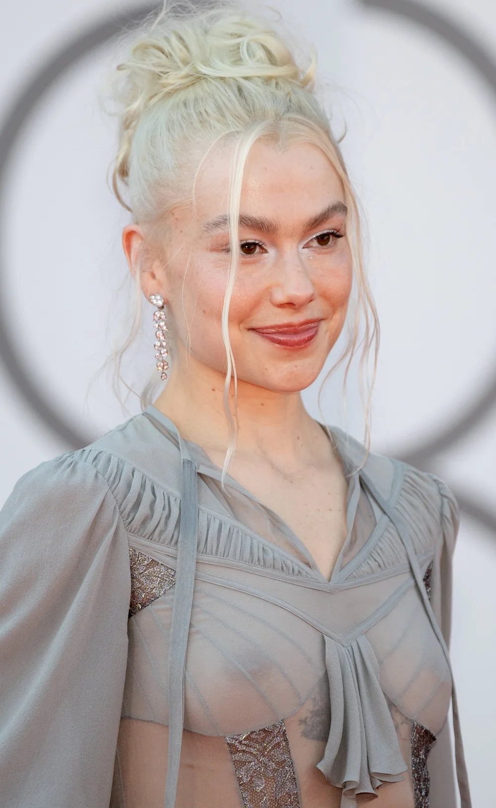 Phoebe-Bridgers-See-Through-Primetime1 A woman with platinum blonde hair in a messy bun, wearing a sheer, gray dress with lace details and dangling earrings, smiles at an event.
