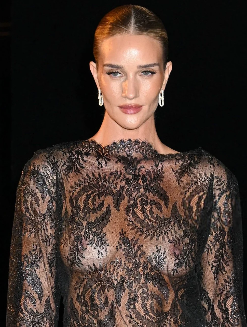 RosieHuntingtonWhiteleySeeThroughYSL26PFWPic2 Close up of woman in sheer lace dress against a black background. She wears makeup and her breasts are visible.