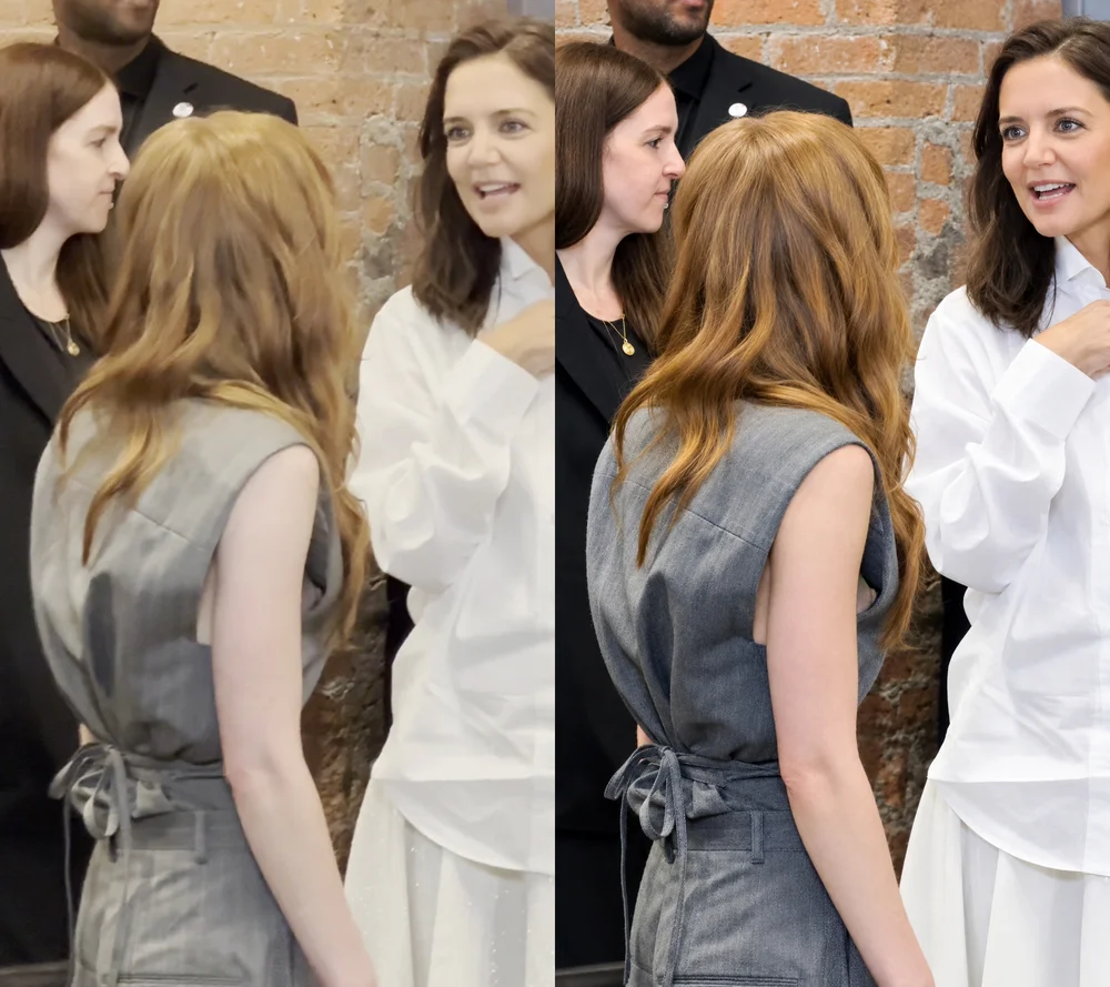 SadieSinkNipSlipCalvinKlein Two women stand facing each other, one with long red hair in a gray dress and the other with brown hair in a white blouse, talking in front of a brick wall. Other people are partially visible in the background.