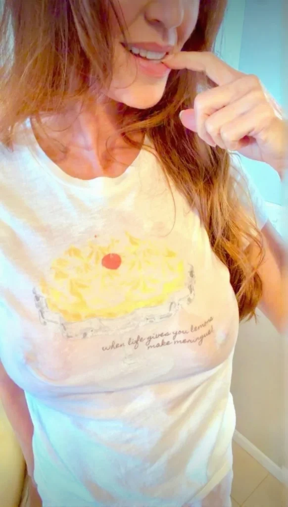 Woman with long brown hair wearing a white t-shirt with a lemon meringue pie and the text when life gives you lemons, make meringue. She is touching her lip and smiling.