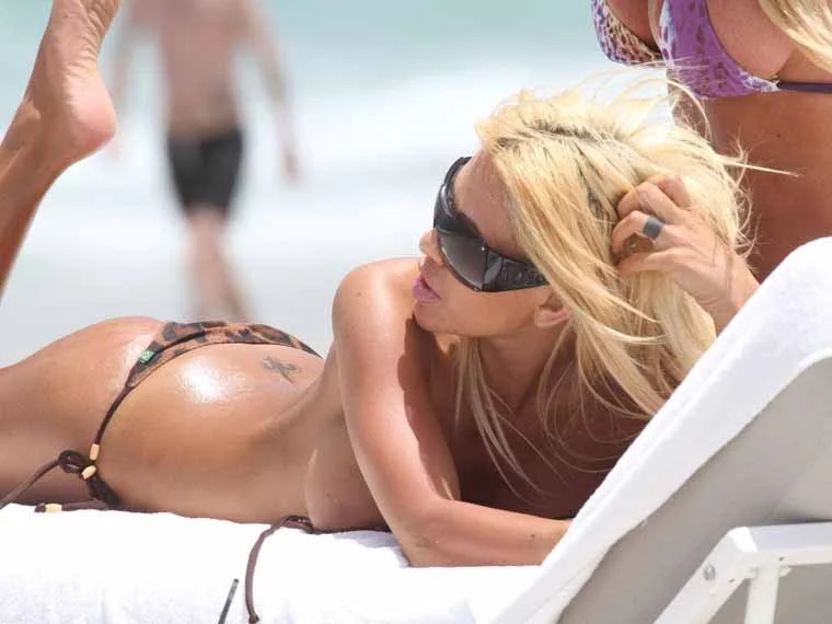 A woman with long blonde hair and sunglasses reclines on a lounge chair at the beach, wearing a bikini. Other beachgoers and the ocean are visible in the background.