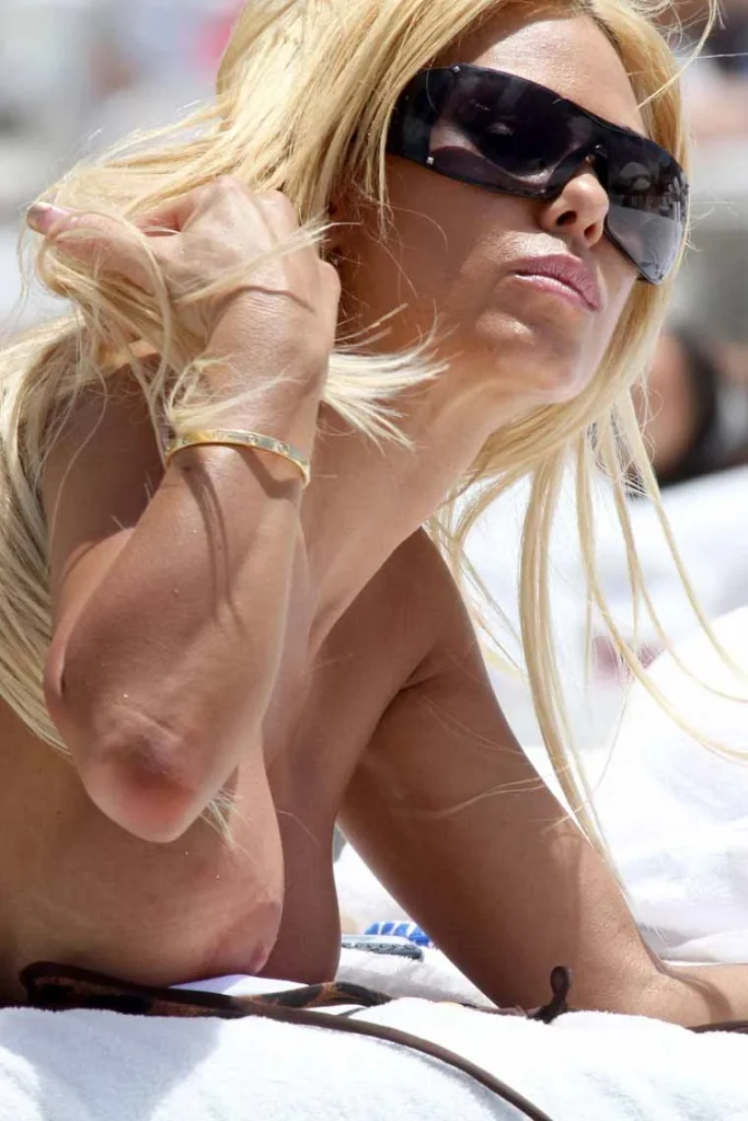 A woman with long blonde hair wearing large black sunglasses is lying on a white towel outdoors, looking upward and adjusting her hair.