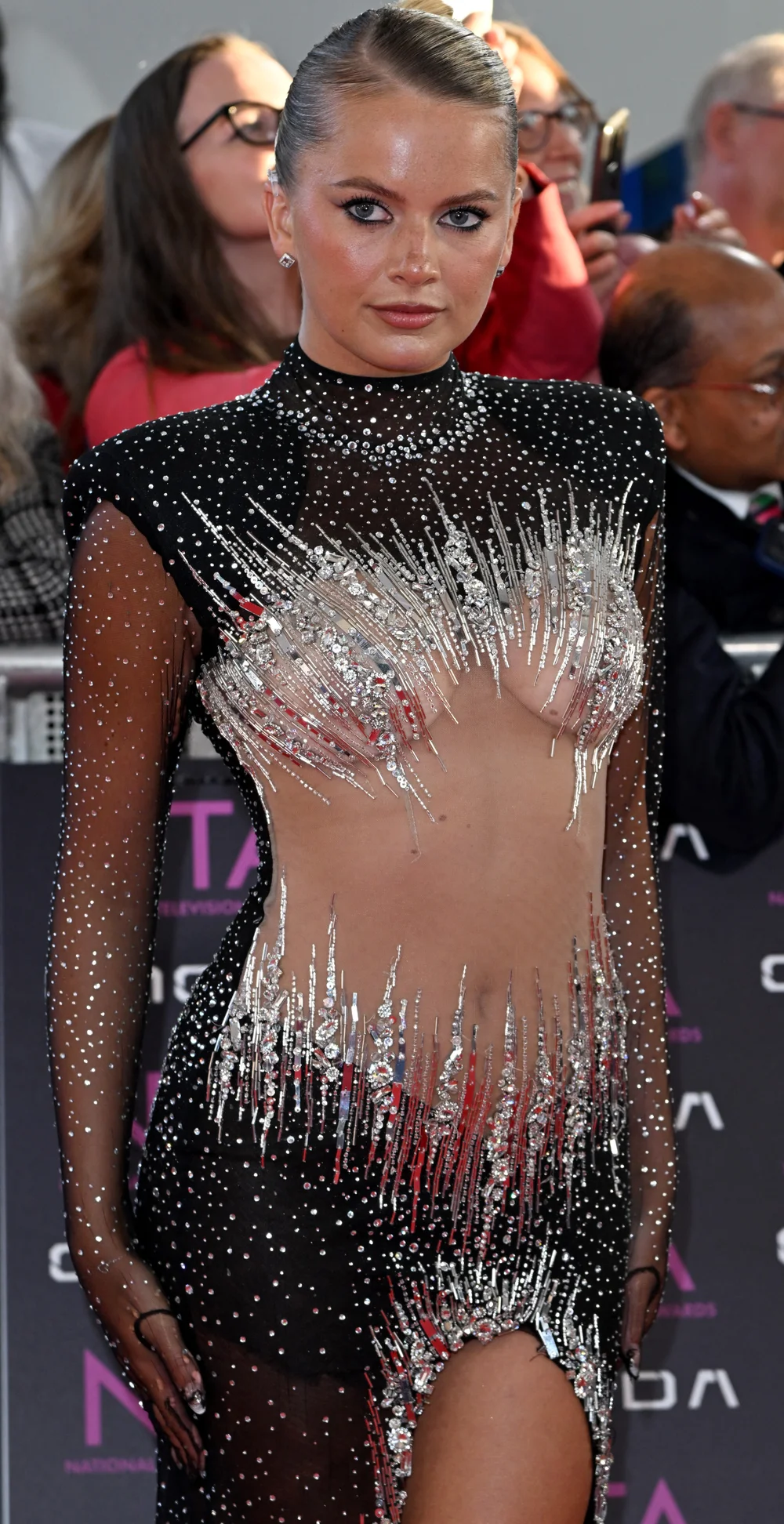 TashaGhouri2026NTAsPic A woman wearing a sheer black and silver sequined dress poses on a red carpet, with people and photographers seen in the blurred background.