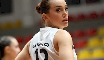 A female volleyball player wearing a white jersey with the number 12 and the name K.Y. GÜLÇİ looks over her shoulder on an indoor court.