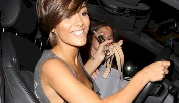 A smiling woman with short brown hair is sitting in the drivers seat of a car, holding a steering wheel. Another person in the passenger seat is holding up an object and partially obscured.