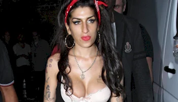 Woman with long dark hair, wearing a red headband, hoop earrings, necklace, and a low-cut top, walks outdoors at night with two men nearby. She has tattoos on her arms and a focused expression.